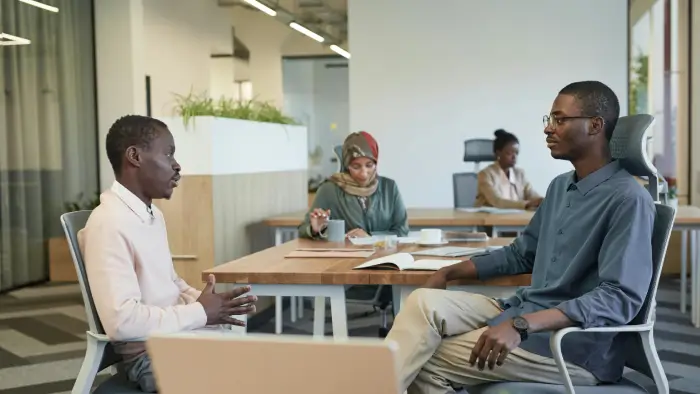 Two men talking in an office with two women working in the background
